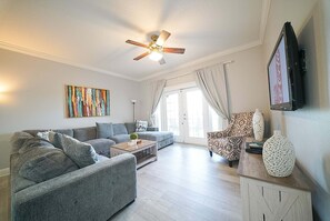 Una sala de estar con sofá, una mesa de centro, un sillón estampado, un ventilador de techo y un televisor montado en la pared.