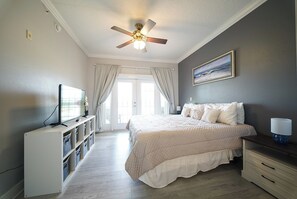 Ein Schlafzimmer mit einem Bett, einem Deckenventilator, einem Fernseher und einem Fenster mit Vorhängen.