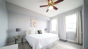 A bedroom with a bed, a ceiling fan, a bedside table, a lamp, a rug, and a window with curtains. - Sunset Greens Retreat at Reunion-7603 (Kissimmee)