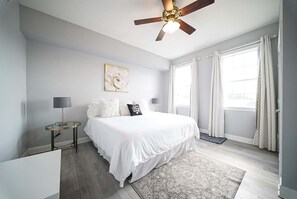 Un dormitorio con cama, ventilador de techo, mesita de noche, lámpara, alfombra y ventana con cortinas.