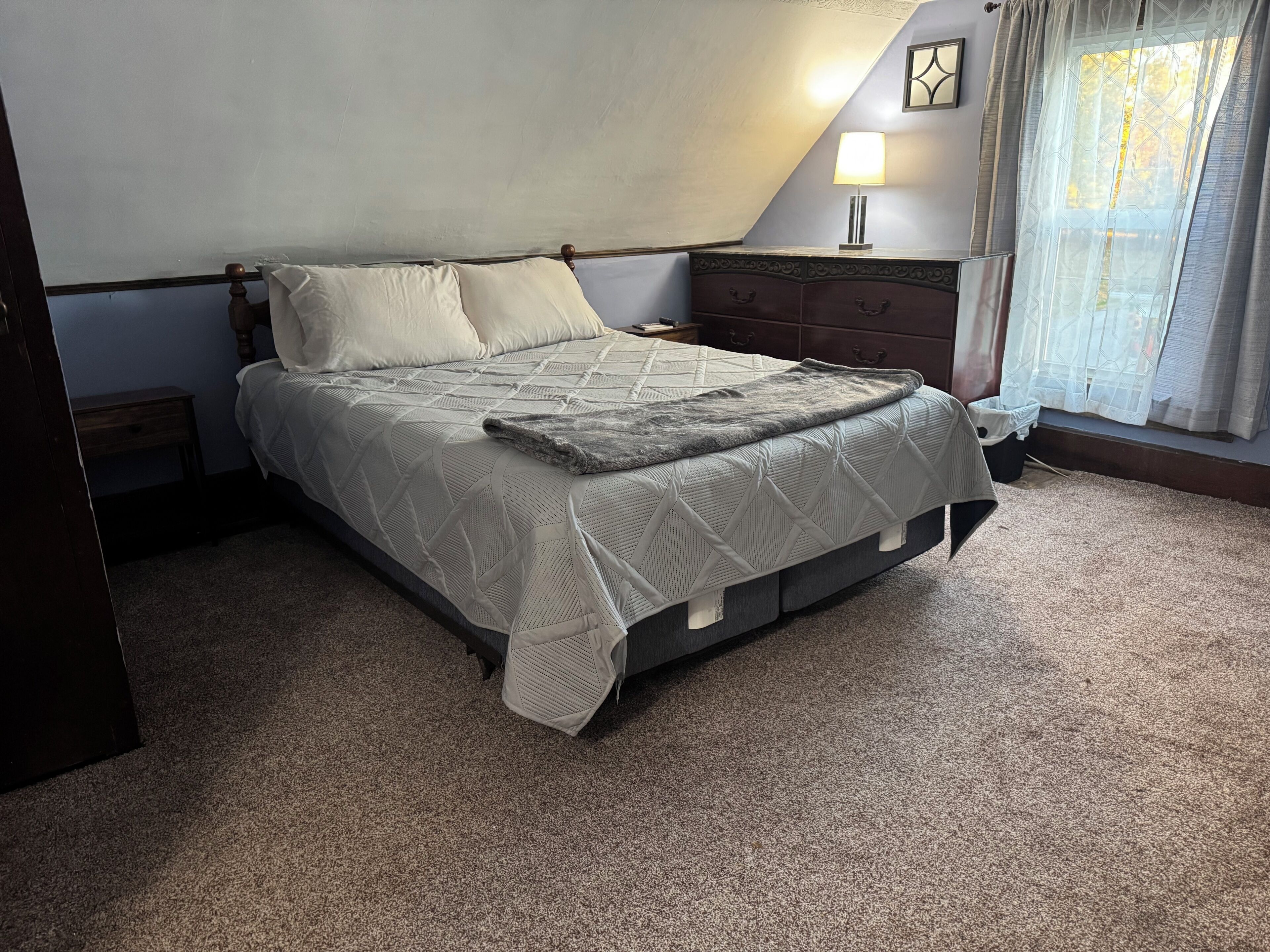 A bedroom with a bed, a nightstand, a lamp, a window with curtains, and a wall-mounted picture.