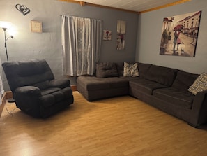 Een woonkamer met een modulaire bank, een zwarte fauteuil, een vloerlamp, een raam met dunne gordijnen en een schilderij van een stel op straat.