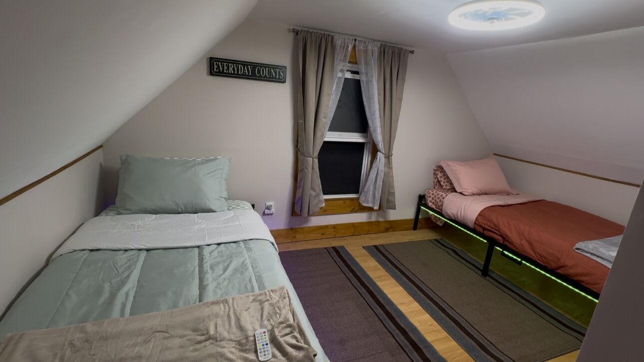 Une petite pièce triangulaire, avec deux lits, une fenêtre avec des rideaux, un panneau sur le mur et un luminaire au plafond.