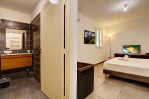 Hotellihuone, jossa on kylpyhuone, sänky, yöpöytä, televisio ja taulu seinällä.
