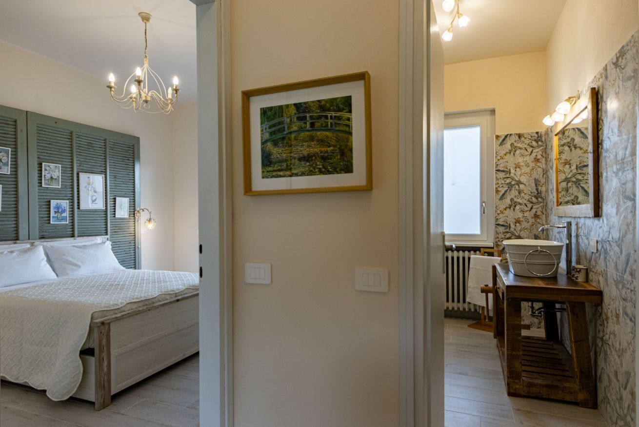 A bedroom with a bed, a chandelier, a framed picture on the wall, and a bathroom with a sink and mirror.