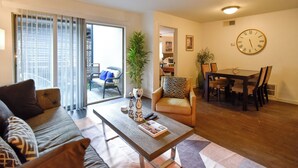 A living room with a sofa, coffee table, dining table, and chairs. There is a potted plant, a clock on the wall, and a sliding glass door leading to a balcony. - The Hidden Gem of Dallas (Dallas)