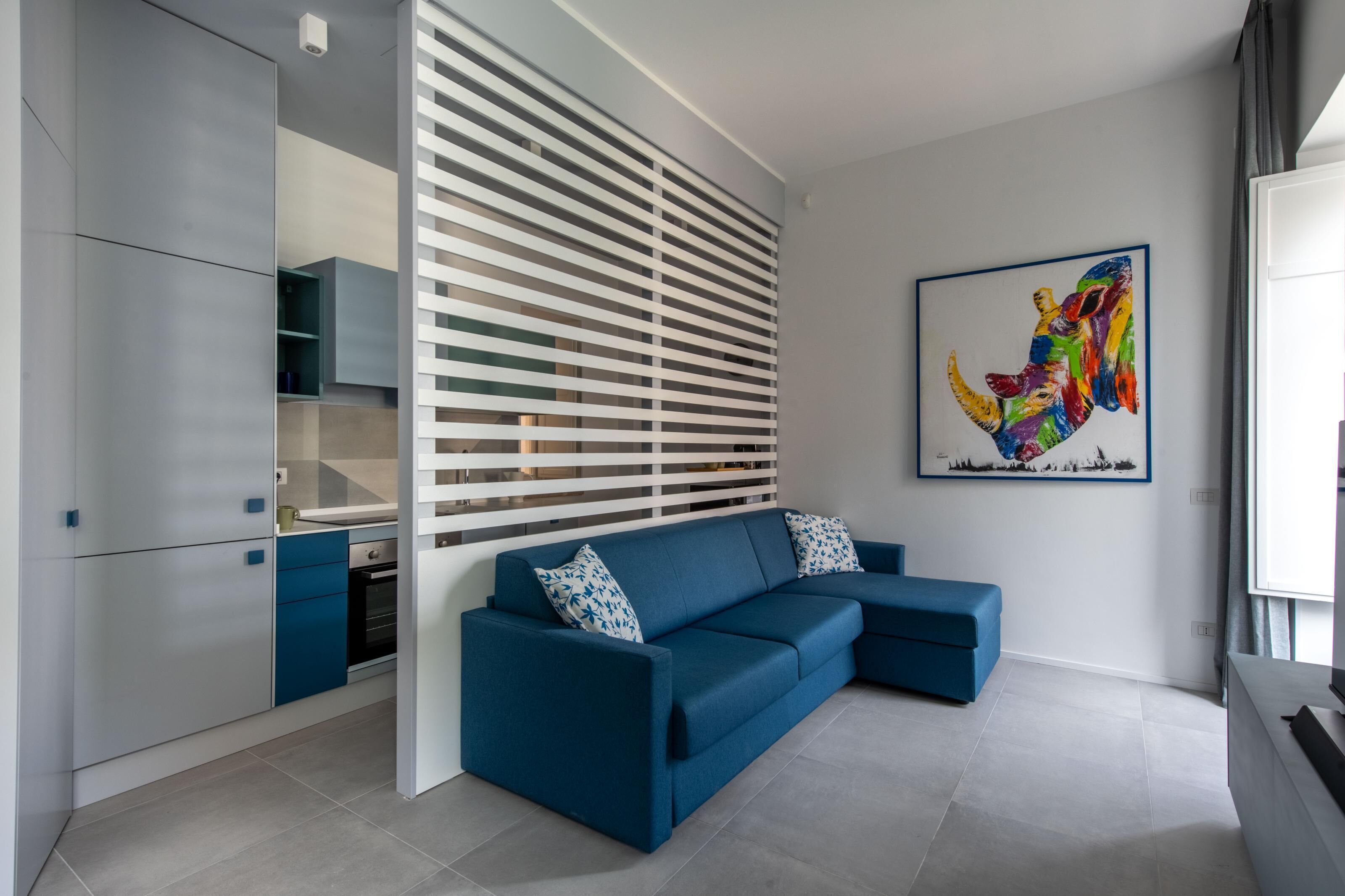 Un salon moderne avec un canapé bleu, un coin cuisine et une œuvre d’art colorée accrochée au mur.