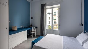 Une chambre à coucher comprenant un lit, un bureau, une chaise et une fenêtre donnant sur des immeubles.