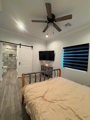Ein Schlafzimmer mit Bett, Deckenventilator, Fernseher und einem Fenster mit Jalousien.