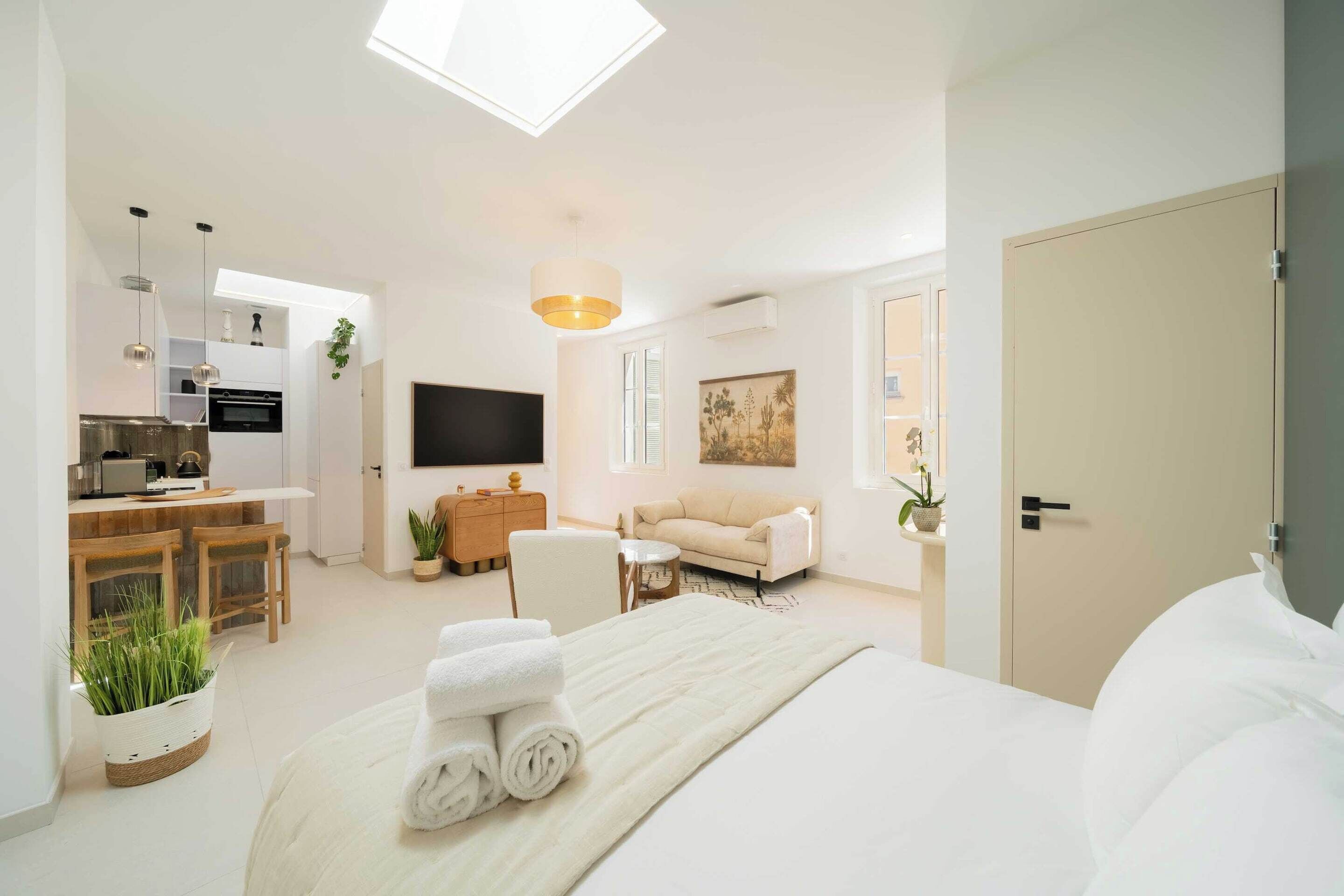 A modern hotel room with a large bed, a flat-screen TV, a sofa, a dining area, and a kitchen.