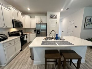 A modern kitchen with white cabinetry, stainless steel appliances, a central island with stools, and a large window. - Modern 3BR Coastal Getaway Near the Beaches of 30A. (Santa Rosa Beach)