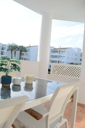 Ein Balkon mit Glastisch, weißen Stühlen, einem Blumentopf und Blick auf weiße Gebäude und Palmen.