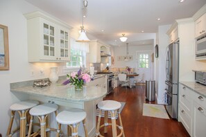 Una cocina con armarios blancos, una isla central con taburetes, electrodomésticos de acero inoxidable y un comedor con una silla.