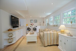 Un área de lavandería con lavadora y secadora, un televisor y un sofá en una habitación luminosa con ventanas amplias.