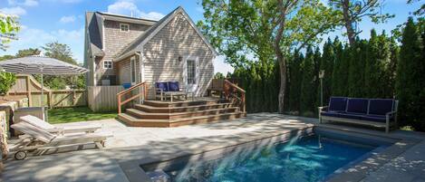A residential swimming pool with a wooden deck, a small house, and a tree.