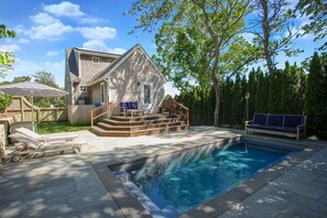 Una piscina residencial con un muelle de madera, una casa pequeña y un árbol.
