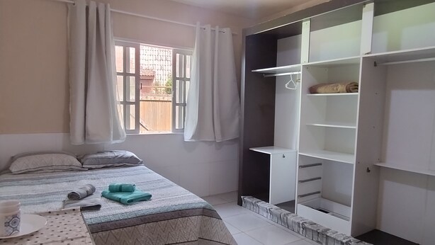 A bedroom with a bed, a window with curtains, a bedside table with a cup, and a wardrobe.