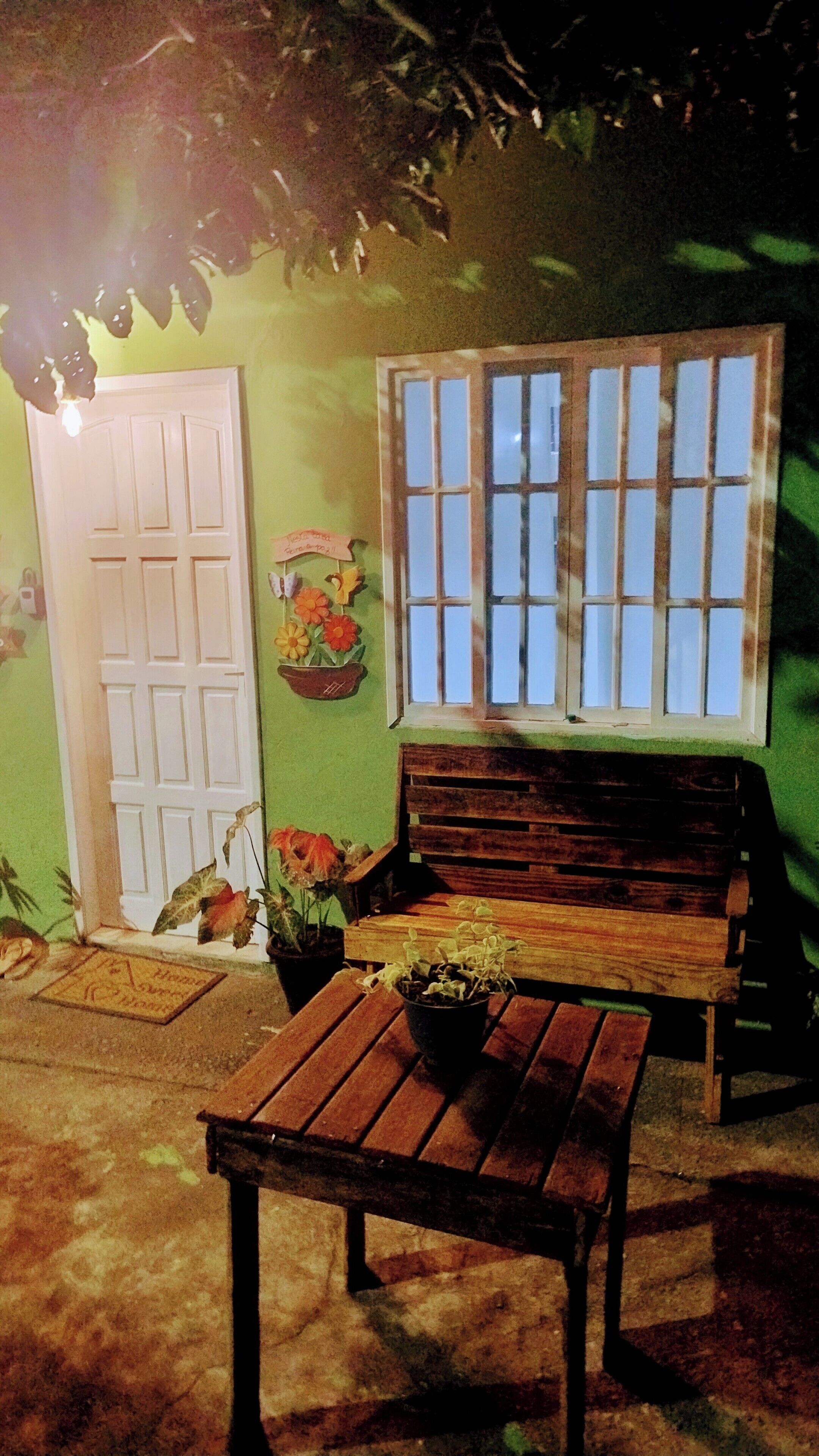 Um banco de madeira, uma mesa, um vaso de planta, uma porta e uma janela com vista.