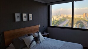 Ett modernt hotellrum med en stor säng, ett träbeklätt sänggavel, ett nattduksbord och utsikt över en stadspanorama genom ett stort fönster.