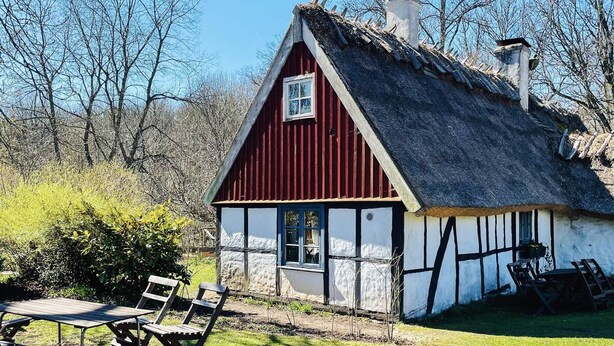 Een vakantiehuisje met rieten dak, rode gevel en witte houten vakwerkbekleding, gelegen in een gazon met bomen.
