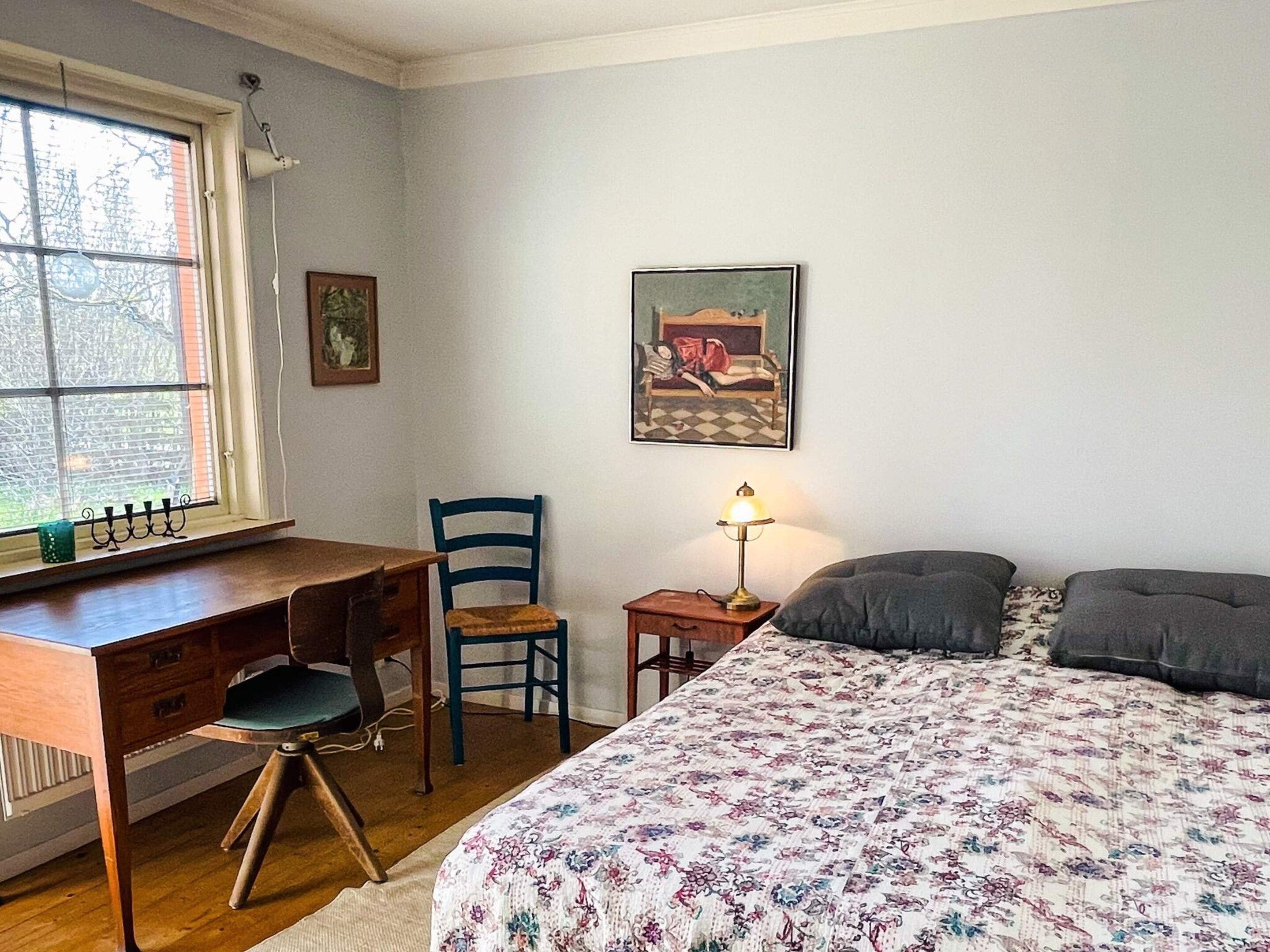 A bedroom with a bed, a desk, a chair, a bedside table with a lamp, and a window with blinds.