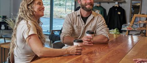 A wooden table with two people holding coffee cups, a window with a view of trees, and a shelf with hats and a framed picture.