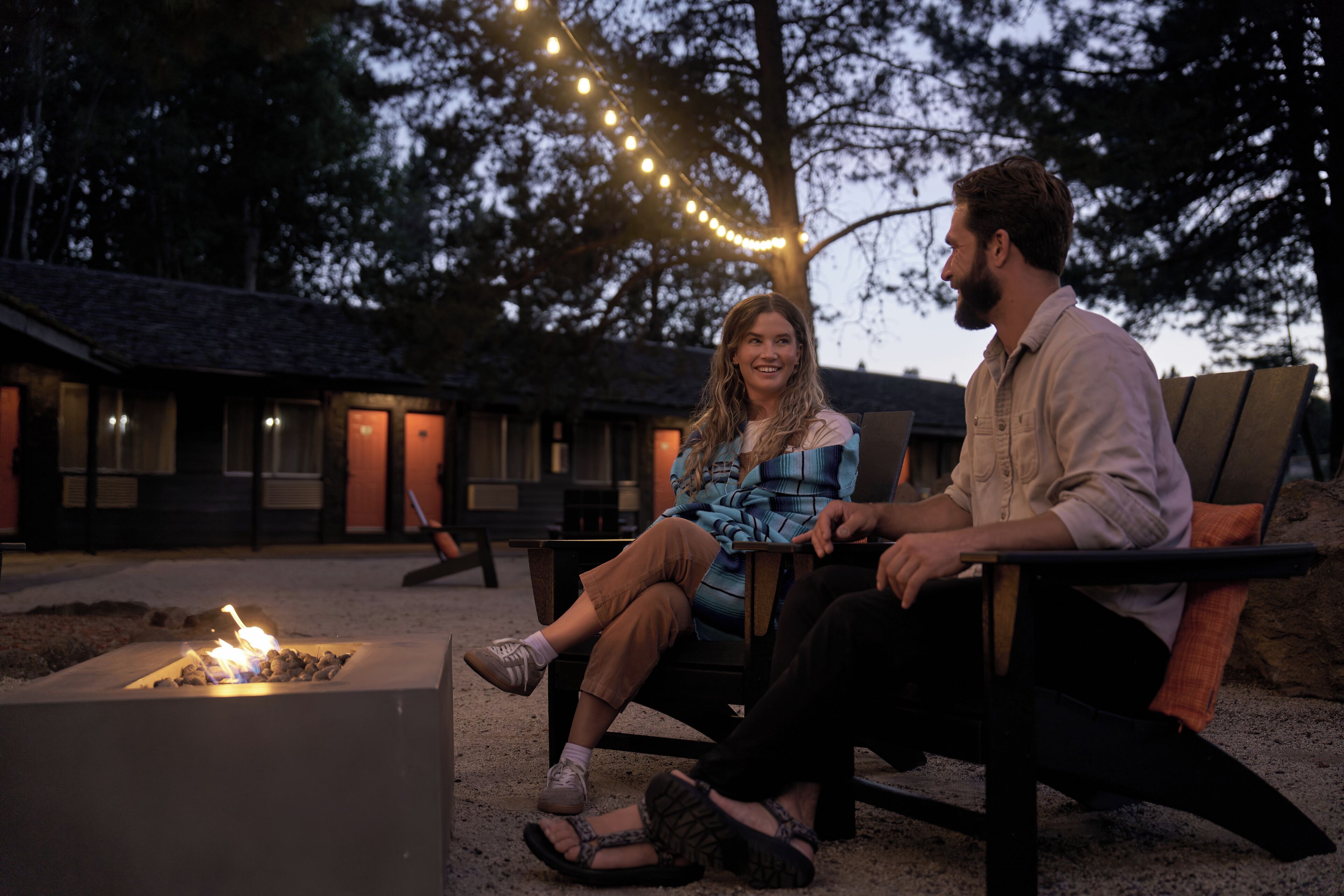 Two people sitting on outdoor chairs near a fire pit, with string lights overhead and a building in the background.
