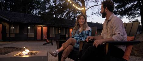 Two people sitting on outdoor chairs near a fire pit, with string lights overhead and a building in the background.