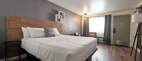 A hotel room with a large bed, a wooden headboard, a bedside table, a chair, a lamp, and a door.