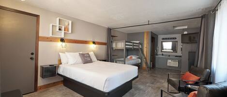 A hotel room with a bed, a desk, a chair, a wardrobe, a mirror, and a kitchenette.