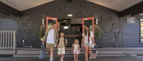 A family of five at a building entrance with a stone facade and a sign indicating amenities like a front desk, lounge, cafe, and general store.