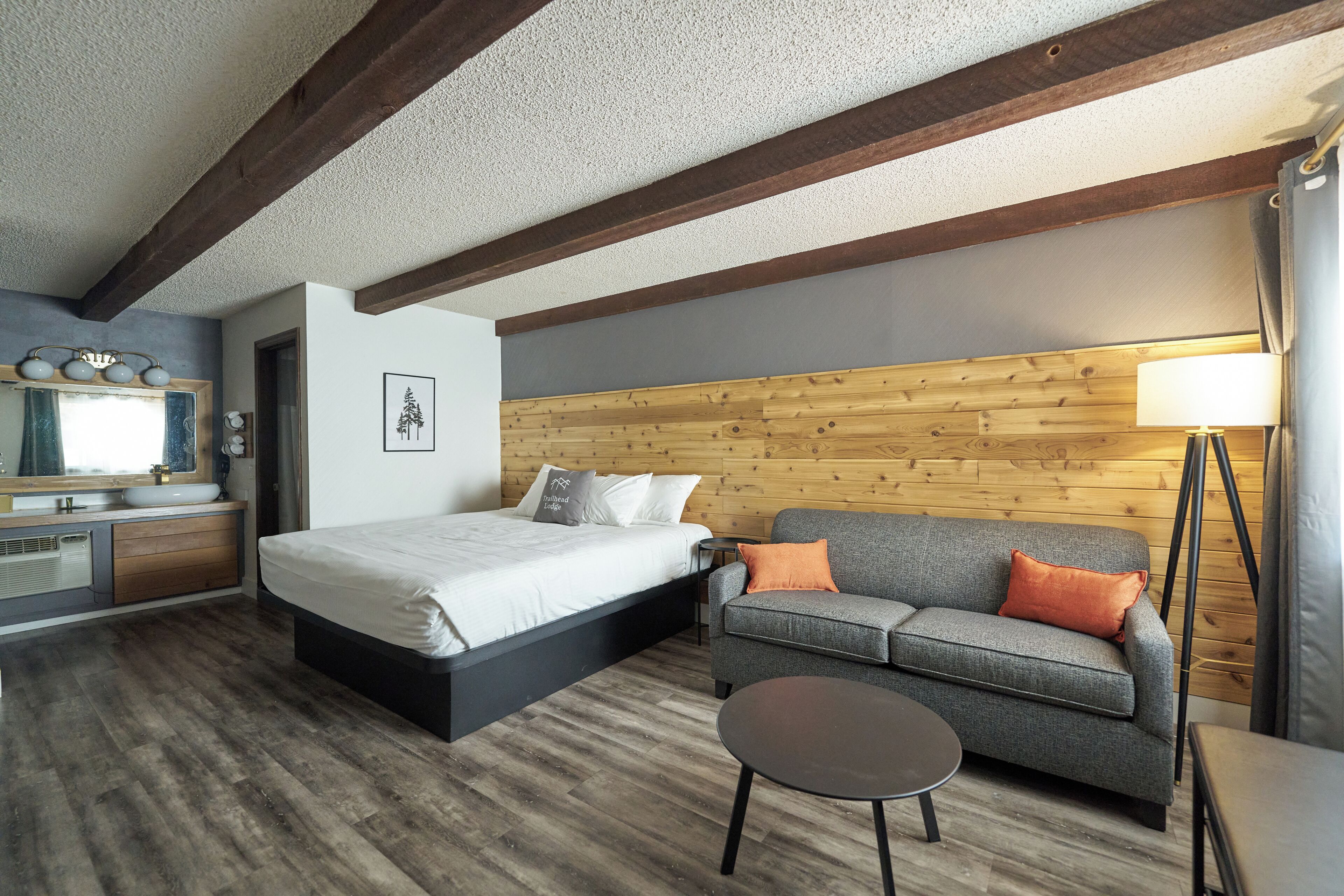 A bedroom with a bed, a sofa, a small table, a lamp, and a wooden wall.