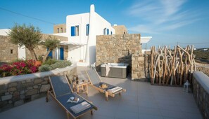 A white building with blue accents, stone walls, and a hot tub. There are lounge chairs and a book on one of them. - Suite With Jacuzzi & Sea View (Mykonos)