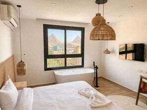 A modern bedroom with a large window, a bathtub, and a view of pyramids. - Luxury panoramic GEM and Pyramids view apartment (Giza Governorate)