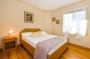 Ein Schlafzimmer mit einem hölzernen Bett, einem Nachttisch mit Lampe, einem Fenster mit leichtem Vorhang und gerahmten Bildern an der Wand.