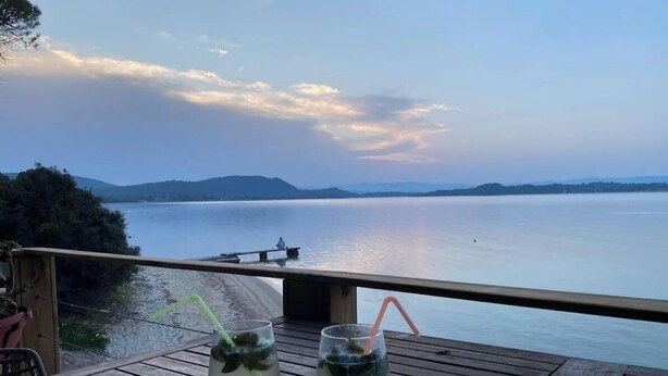 A wooden deck with two glasses of iced drinks, a bench, and a view of a calm lake with distant hills. - Beachfront Private house in Vourvourou, Chalkidiki with amazing view (Vourvourou)