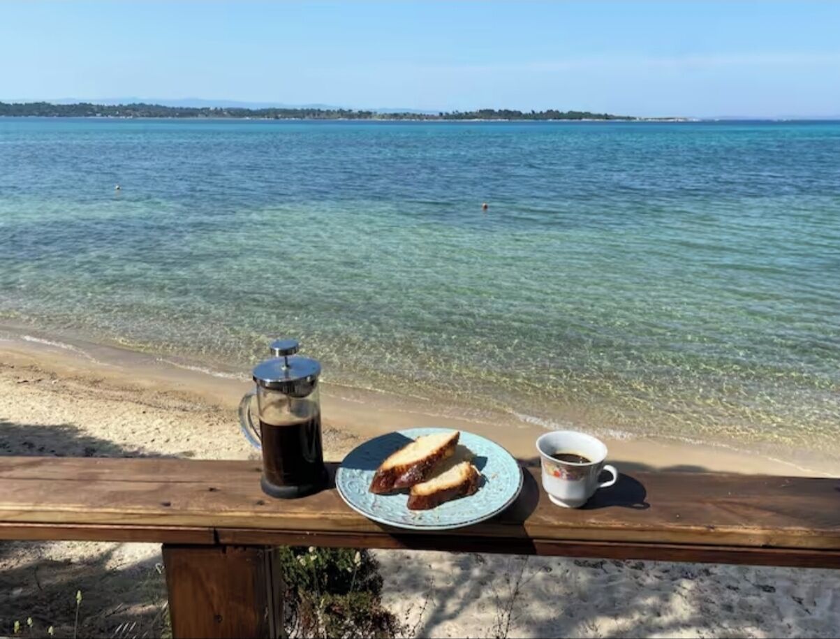 Un banco de madera con un plato de comida y una taza de café, con vista a un mar azul y despejado.