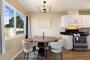 A modern kitchen with white cabinets, a round dining table with chairs, a chandelier, and a view of the outside. - Oceanview 2BR 1Bath Balcony Sunset Views (Pacifica)