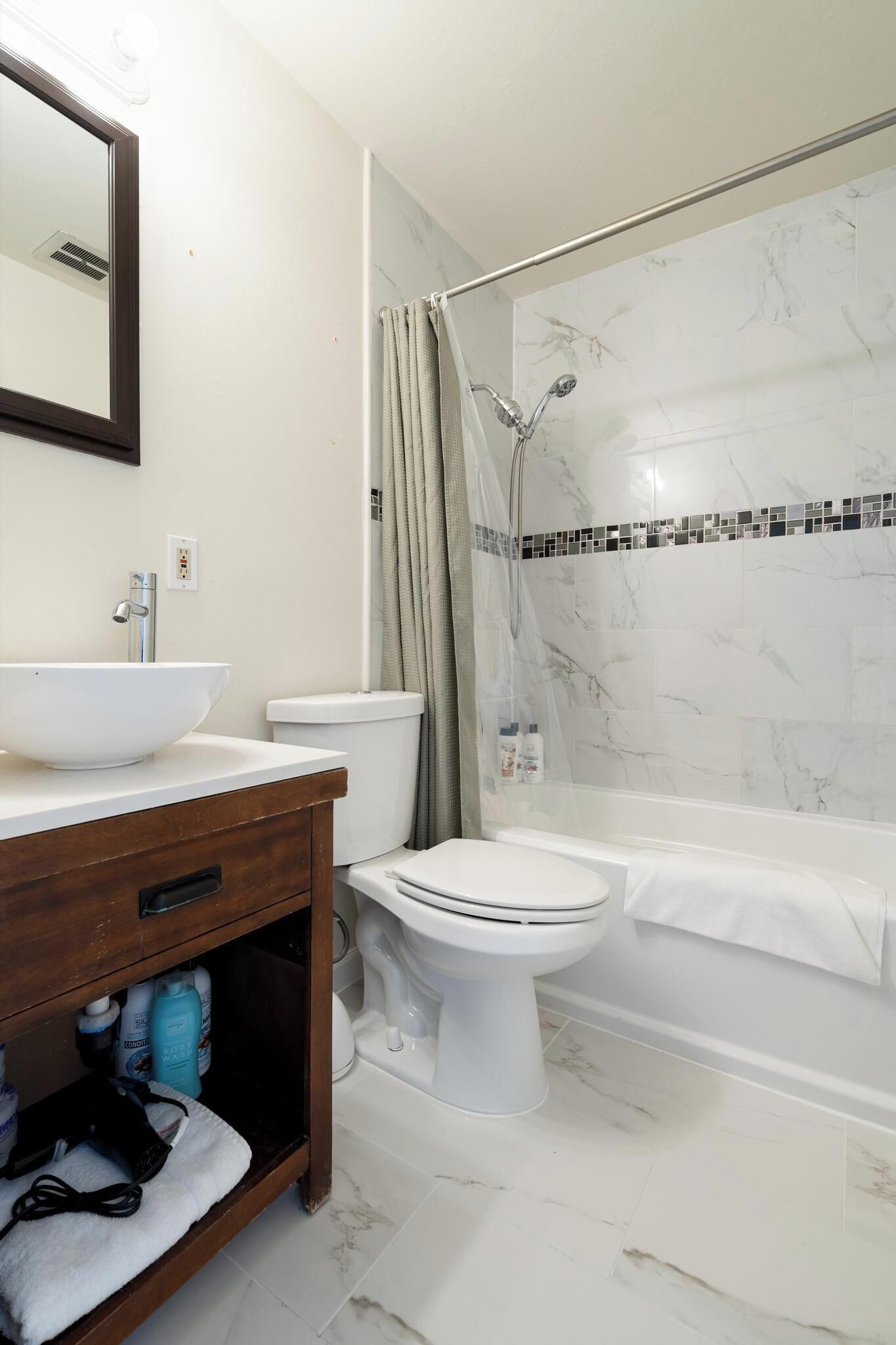 A bathroom with a white sink, toilet, and bathtub, marble tiles, and a wooden vanity.
