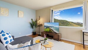 A modern living room with a large TV, a sofa, a coffee table, and a view of a coastal landscape. - Oceanview 2BR 1Bath Balcony Sunset Views (Pacifica)