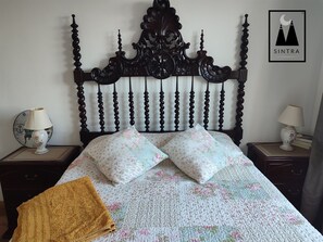 Ein Bett mit dunklem Kopfteil, floralen Kissen und einer Tagesdecke. An den Nachttischen stehen Lampen, und an der Wand hängt ein gerahmtes Bild.