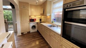 A compact kitchen with wooden cabinets, a washing machine, a microwave, and a built-in oven. - Room-cosy Single Room Near CBS Arena and (Coventry)