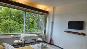 A hotel room with a bed, a TV mounted on the wall, a small table with chairs, and a window with a view of trees. - Studio with lake view (San Carlos de Bariloche)