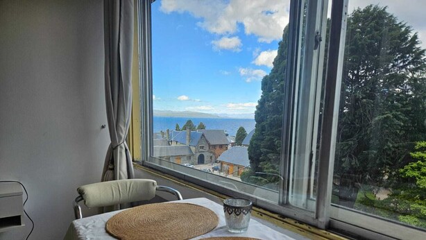 A room with a table set for two, a view of a coastal town, and a window with curtains. - Studio with lake view (San Carlos de Bariloche)