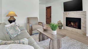 Ett vardagsrum med en soffa, fåtölj, soffbord, öppen spis, en platt-TV och en lampa.