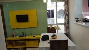 A room with a yellow entertainment unit, a television mounted on the wall, a table with a potted plant, and a window with curtains.
