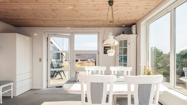 Una habitación con techo de madera, muebles blancos, una mesa de comedor con sillas y ventanas amplias que ofrecen vistas al exterior.
