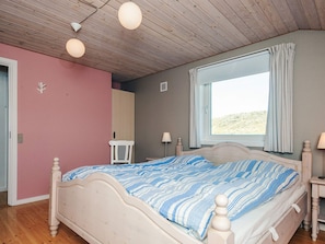 Ein Schlafzimmer mit einem Bett, einem Fenster mit Vorhängen, einem Nachttisch und einem Sessel.