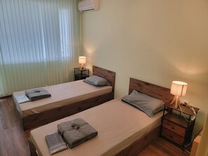 Une chambre double avec deux lits, des tables de chevet avec des lampes, une fenêtre avec des stores et un appareil de climatisation.