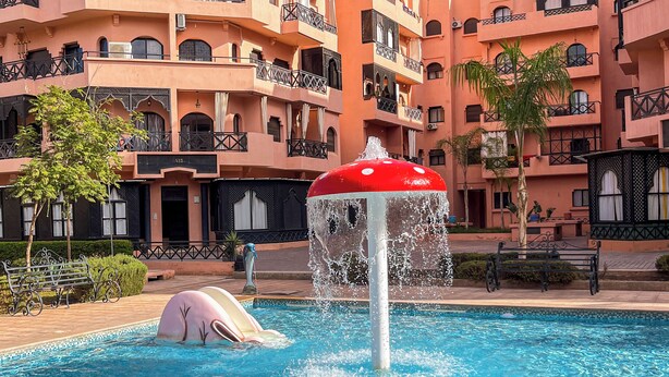 A swimming pool with a water feature, surrounded by multi-story buildings with balconies and a clear blue sky. - Palmeraie fountain view (Marrakech)